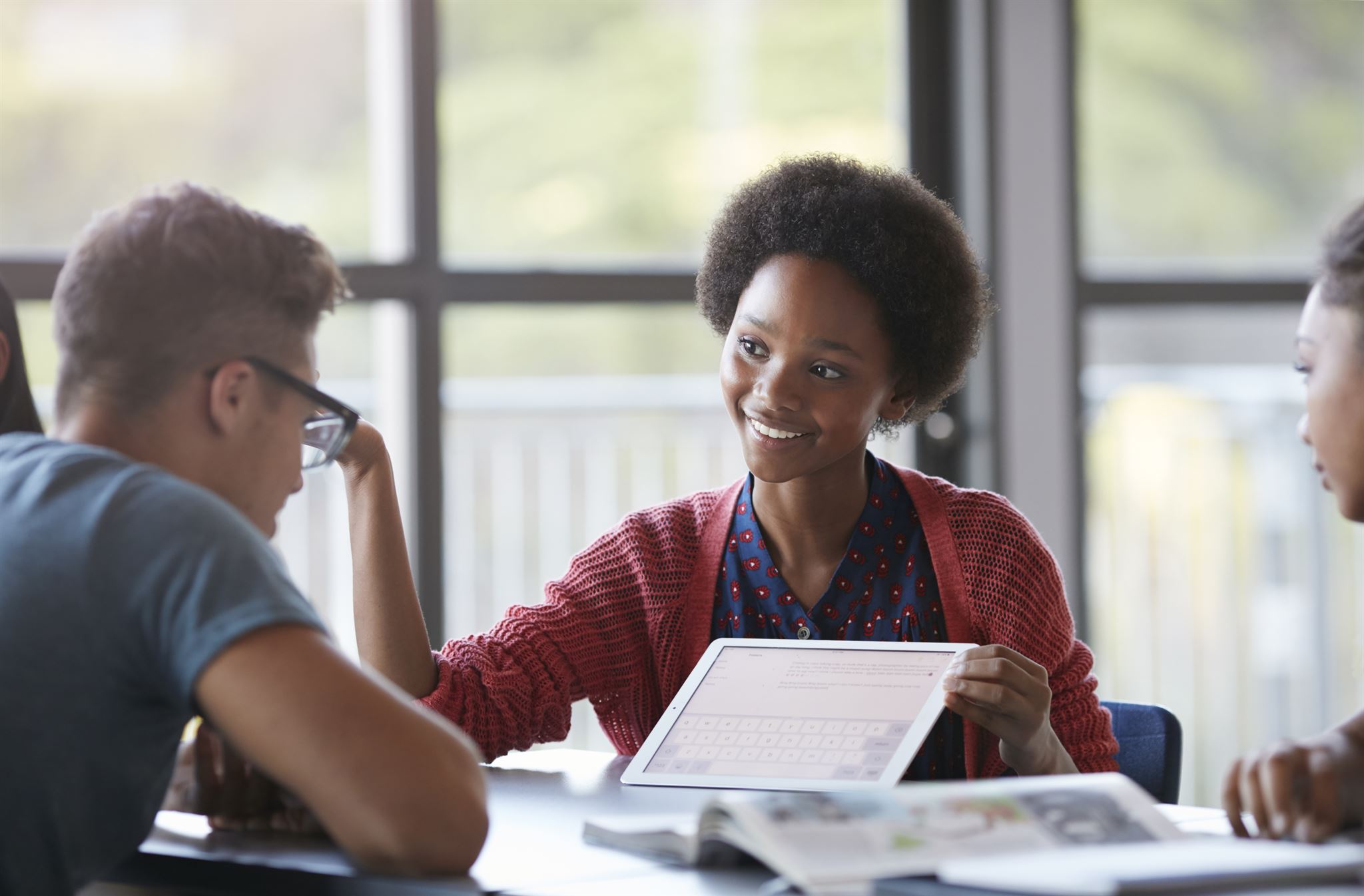 Student-seated-at-table-presenting-on-a-tablet-to-two-other-students